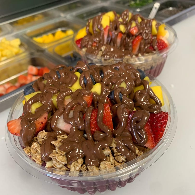 Two fruit bowls topped with granola, strawberries, blueberries, mango, and chocolate drizzle.
